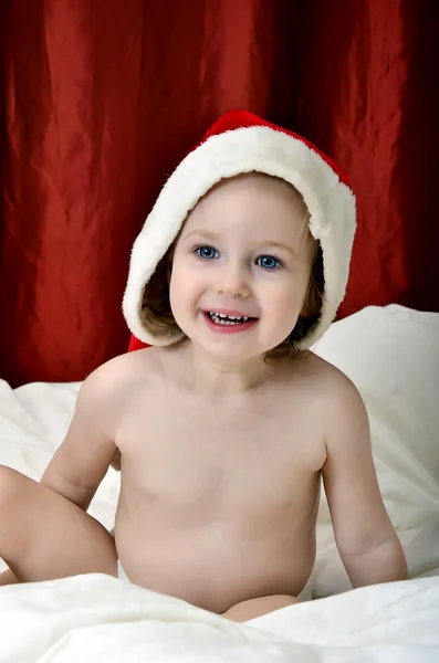 Little girl in a Christmas red hat sitting on a bed — Stock Photo, Image