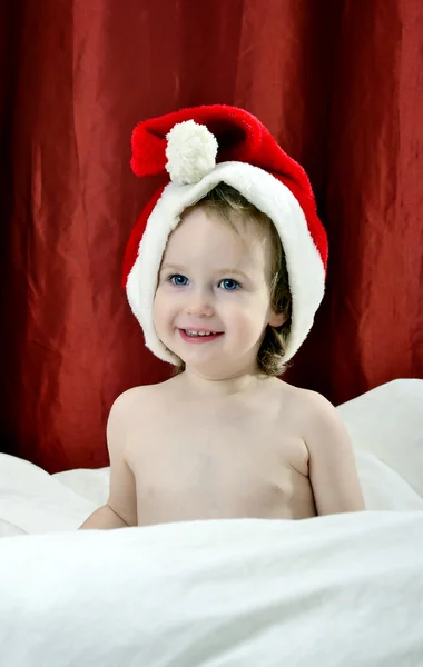 Κοριτσάκι σε ένα χριστουγεννιάτικο κόκκινο καπέλο που κάθεται σε ένα κρεβάτι — Φωτογραφία Αρχείου