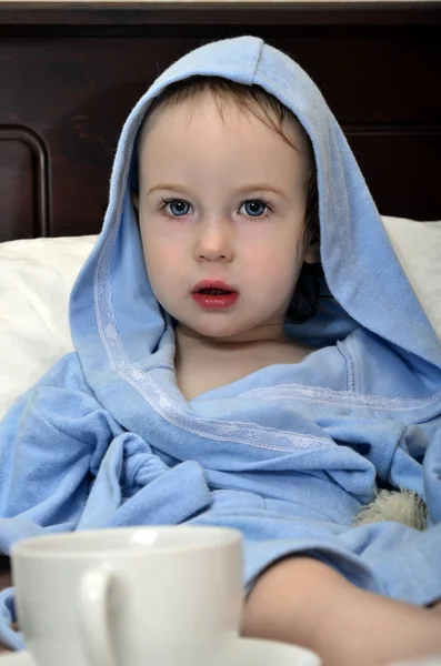 Маленькая девочка в синем халате отдыхает на кровати после душа с чашкой — стоковое фото