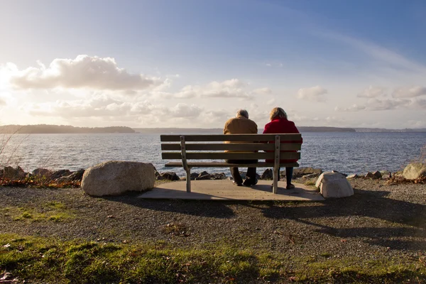 Casal de idosos relaxando juntos no banco do parque de frente para um lago — Fotografia de Stock