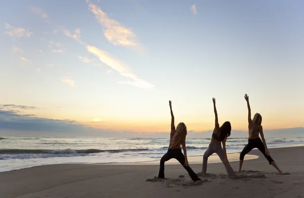 Tři ženy cvičí jógu na pláži při východu nebo západu slunce — Stockfoto
