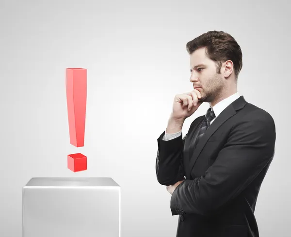 Jovem empresário olha para o ponto de exclamação vermelho em um pedestal branco . — Fotografia de Stock