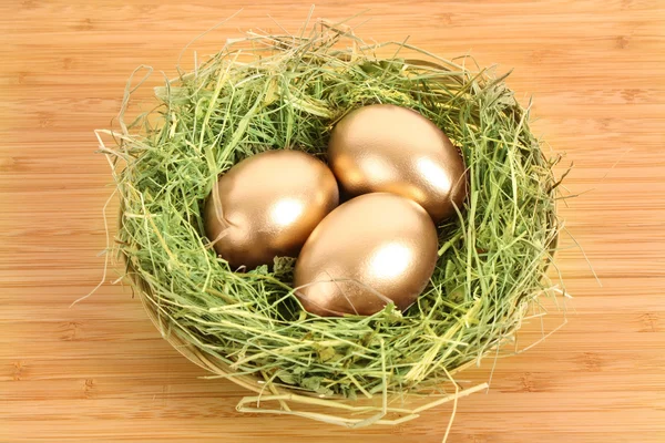 Trzy złote kurze jaja w gnieździe trawiastych na drewnianym stole — Zdjęcie stockowe