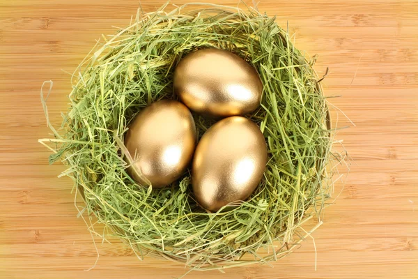 Three golden hen's eggs in the grassy nest on the wooden table — Stock Photo, Image