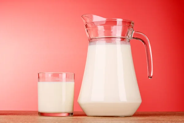Džbán a sklenice s mlékem na červeném pozadí — Stock fotografie