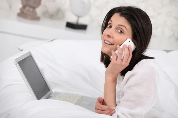 Γυναίκα χρησιμοποιώντας ένα τηλέφωνο και το φορητό υπολογιστή στο υπνοδωμάτιό της — Φωτογραφία Αρχείου