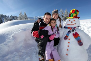 Couple posing with child beside snowman at mountain resort clipart
