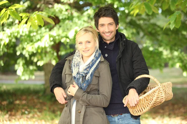 Giovane coppia dietro gli alberi in autunno, uomo che tiene un cesto di vimini — Foto Stock