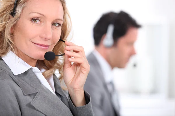 Γυναίκα σε ένα κοστούμι χρησιμοποιώντας ένα ακουστικό με ένα αρσενικό συνάδελφο στο παρασκήνιο — Φωτογραφία Αρχείου