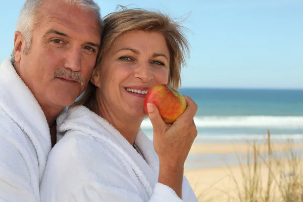 Äldre par i badrockar äta ett äpple på stranden — Stockfoto