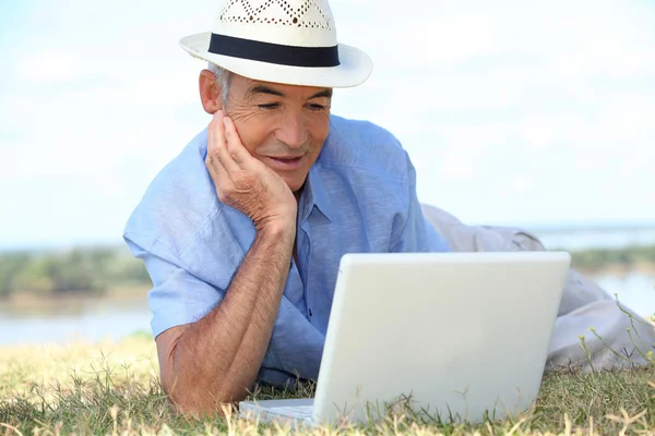 Hombre mayor acostado en la hierba bajo el sol usando una computadora portátil —  Fotos de Stock