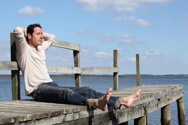 Uomo scalzo seduto su un molo di legno godendo il sole — Foto Stock