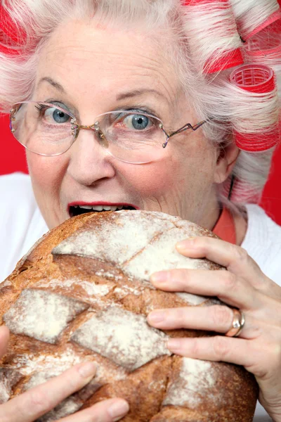 An old lady with hairroller on, biting on a loaf of bread. — Stock Photo, Image