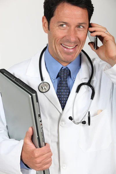 Ein Arzt hält einen Laptop in der Hand und führt ein Gespräch über sein Mobiltelefon. — Stockfoto