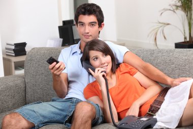 Young couple at home, watching TV and talking on the telephone clipart