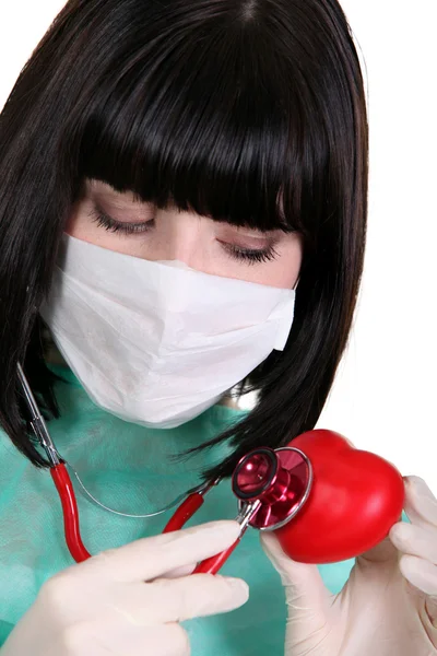 Black-haired nurse with surgical mask using stethoscope on red heart — Stok fotoğraf