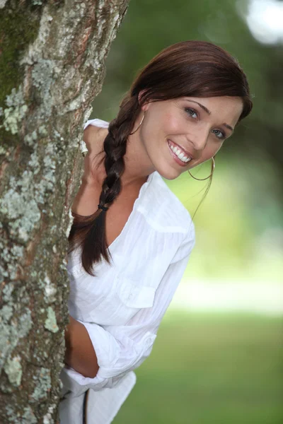 Femme souriante penchée derrière un arbre — Photo