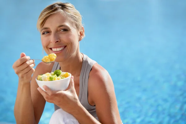 Woman eating fruit cocktail by swimming pool — Stockfoto