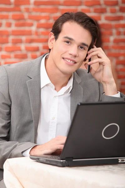 Νεαρός άνδρας, χρησιμοποιώντας ένα φορητό υπολογιστή και το τηλέφωνο σε ένα εστιατόριο — Φωτογραφία Αρχείου