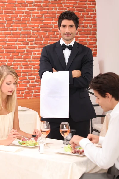Waiter stood by couple enjoying meal Stock Picture
