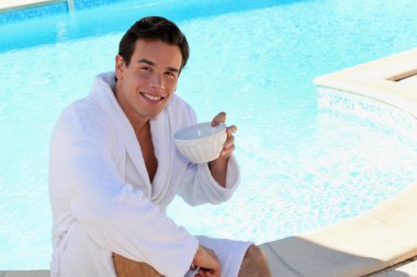 Young man dressed in bathrobe sitting on the edge of a swimming pool and dr clipart