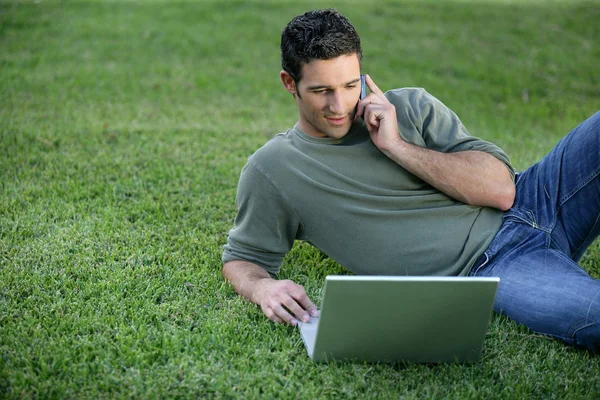 Relajado hombre usando el teléfono y la computadora puesta en la hierba —  Fotos de Stock