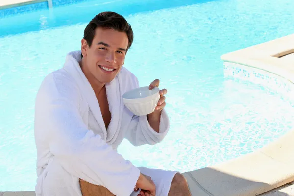 Young man dressed in bathrobe sitting on the edge of a swimming pool and dr — Stock Photo, Image