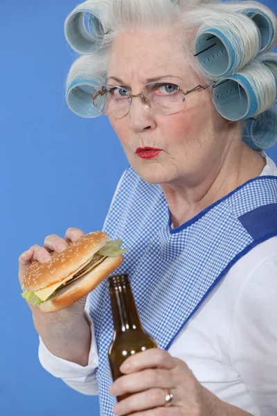 Sénior com rolos no cabelo bebendo cerveja e comendo hambúrguer — Fotografia de Stock
