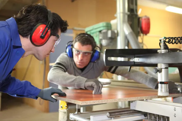 Un trabajador experimentado mostrando a un aprendiz cómo cortar un trozo de madera usin —  Fotos de Stock