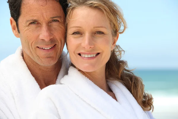 En man och en kvinna klädd i morgonrockar och ler mot oss på en strand. — Stockfoto