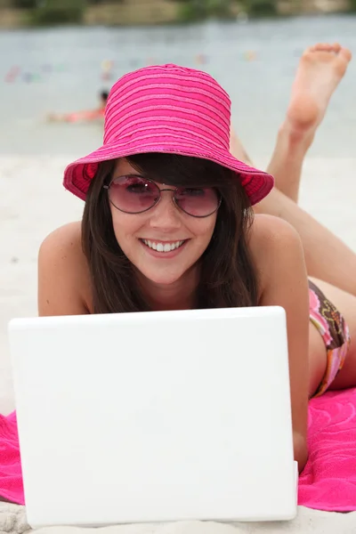 Jovem mulher em um chapéu rosa brilhante usando um computador portátil na praia — Fotografia de Stock