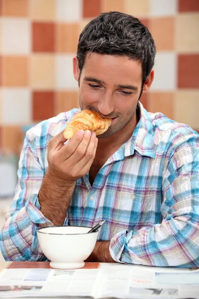 Улыбающийся мужчина ест круассан на завтрак с журналом — стоковое фото
