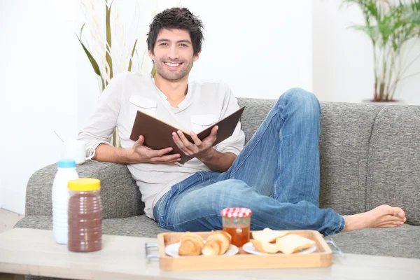 Ο άνθρωπος που κάθεται σε έναν καναπέ τρώει πρωινό και διαβάζοντας ένα βιβλίο — Φωτογραφία Αρχείου