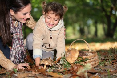 A mother and her little girl picking mushrooms in the forest clipart