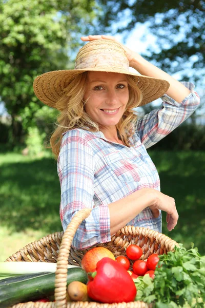 Woman with a hand on her hat and basket of vegetables. — Stock Photo, Image