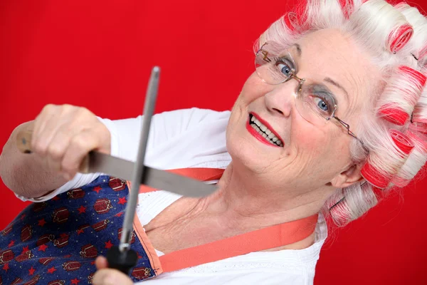 Бабушка с бигуди затачивает ножи на красном фоне — стоковое фото