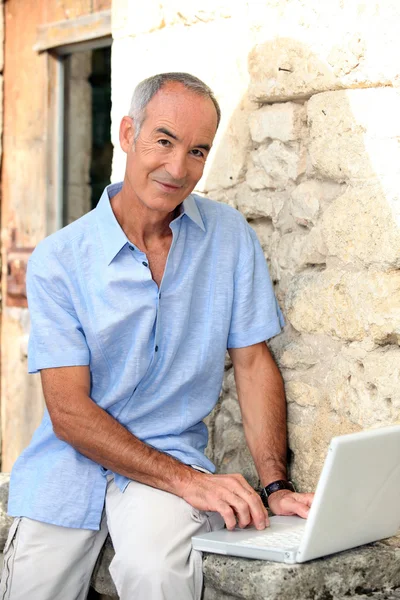 Szare włosy człowiek siedział przez kamienny mur z laptopa — Zdjęcie stockowe