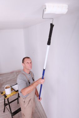 Decorator using a roller extension to paint a ceiling white clipart
