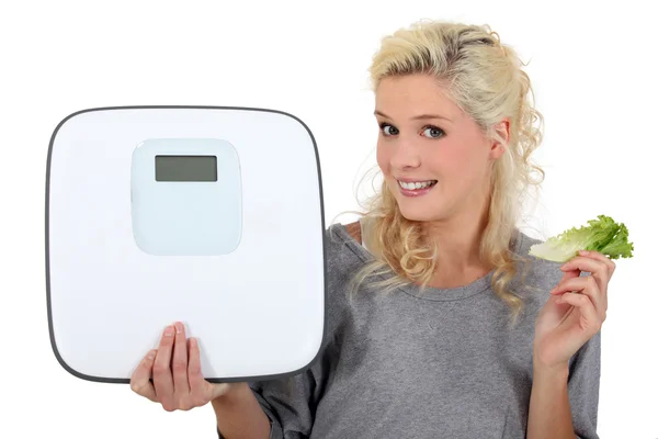 Blonde woman showing a bathroom scale and taking a lettuce leave — Stock Photo, Image