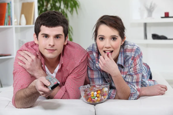 Young couple watching TV with a remote control and bowl of sweets — Stock Photo, Image