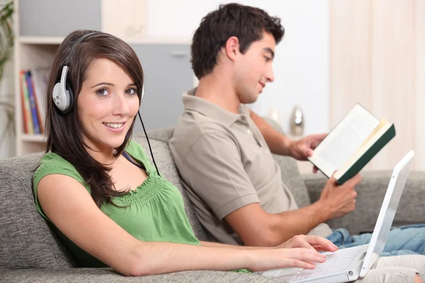 Woman swearing headphones and typing on laptop whilst boyfriend reads — Stock Photo, Image