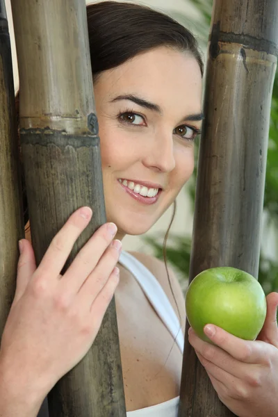 Portrait of smiling brunette behind bamboos with green apple — Stock Photo, Image