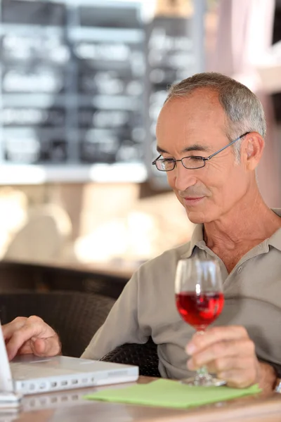 Homme aux cheveux gris utilisant un ordinateur portable tout en buvant un verre de vin — Photo