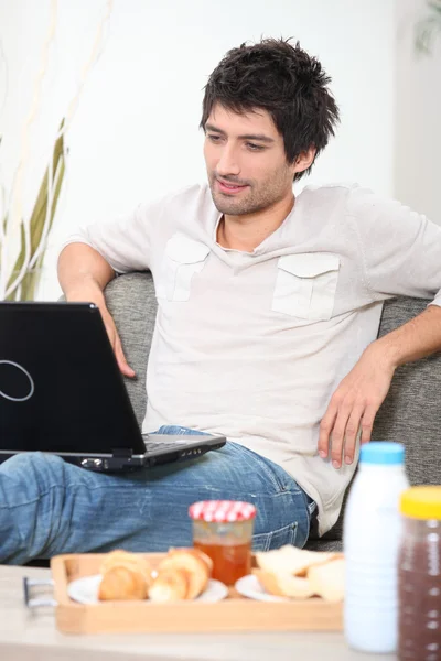 Άνθρωπος χρησιμοποιώντας ένα φορητό υπολογιστή στο δωμάτιό του μέτωπο με πρωινό σε ένα δίσκο — Φωτογραφία Αρχείου