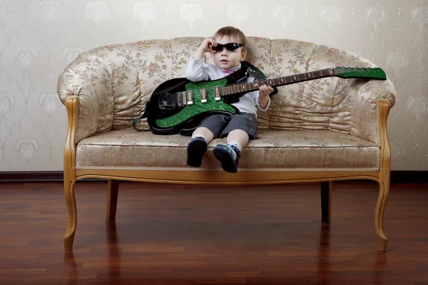 Pequeno menino guitarrista de moda sentado no sofá glamouroso — Fotografia de Stock