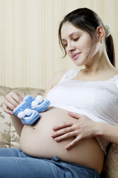 Těhotná žena na gauči s modré botičky v rukou — Stock fotografie