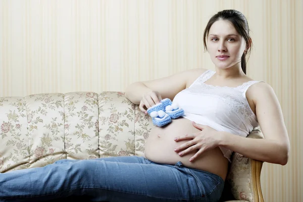 Těhotná žena na gauči s modré botičky v rukou — Stock fotografie