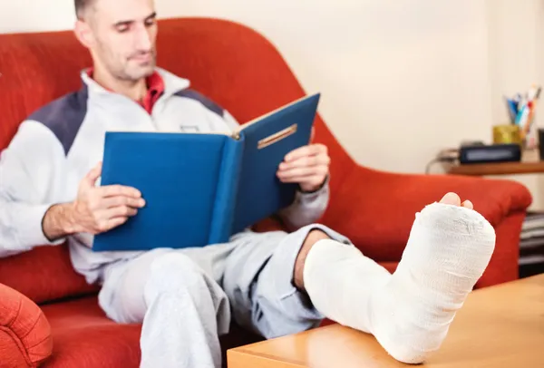 Man with a broken leg on a sofa at home reading book — Stock Photo, Image