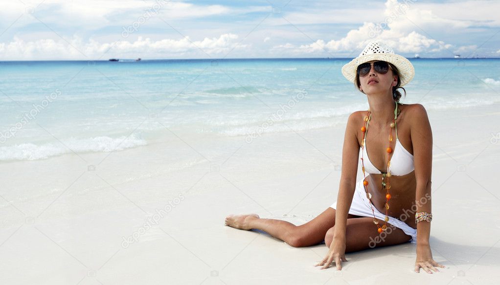 Attracrive girl in a bikini and straw hat sitting on the sand of a tropical