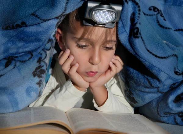 Niño leyendo un libro debajo de las cubiertas con una linterna — Foto de Stock